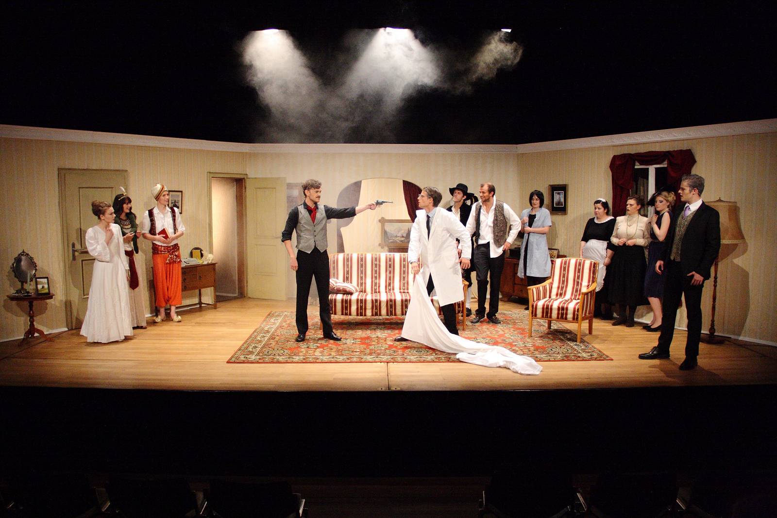 Ein Theaterstück, in dem ein Mann in der Mitte der Bühne eine Waffe auf einen anderen Mann im weißen Kittel richtet. Die Bühne sieht wie ein Wohnzimmer aus, drum herum stehen andere Schauspieler.